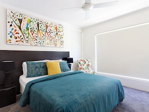 A modern bedroom with a bed, a nightstand, a chair, a ceiling fan, and a large abstract painting on the wall.