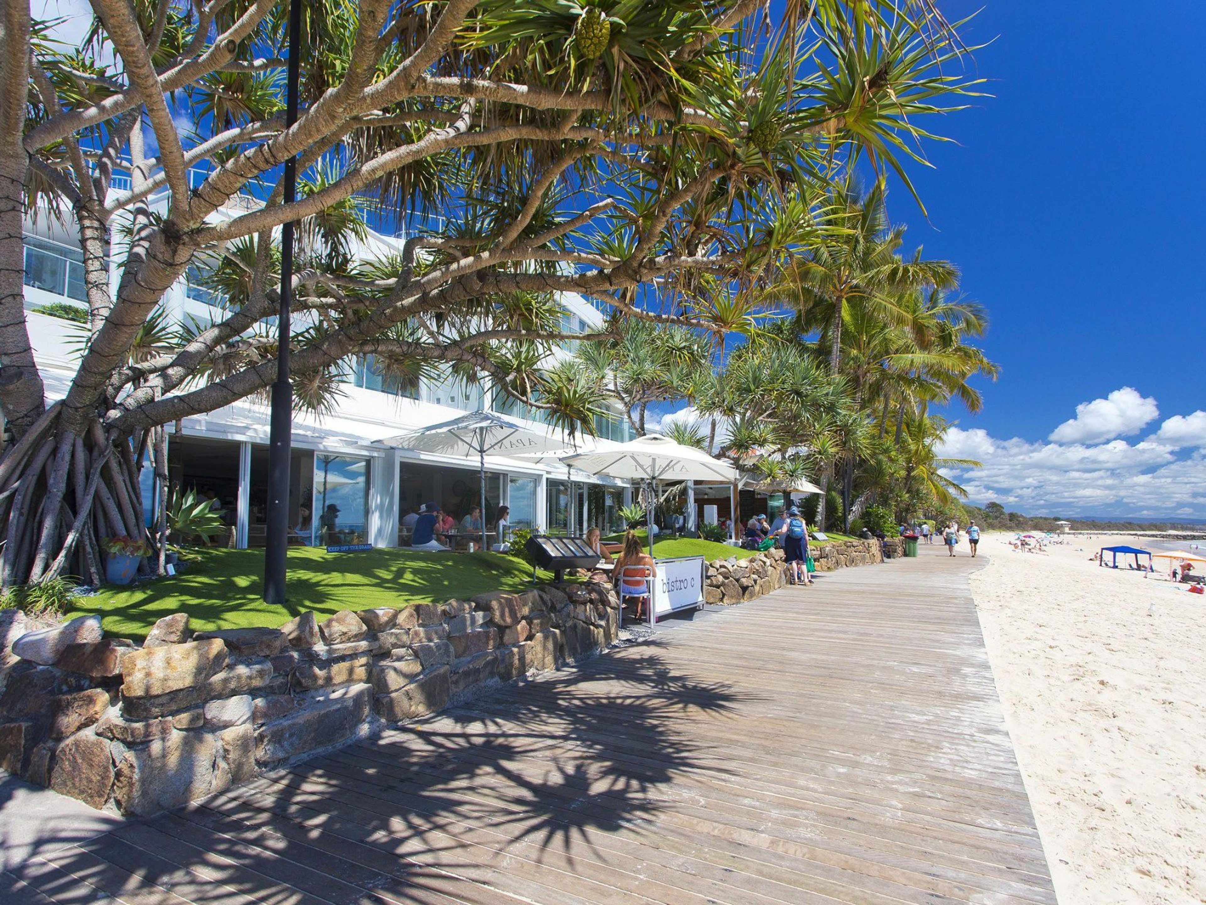 Un hotel sulla spiaggia con posti a sedere all'aperto, una passerella in legno e palme.