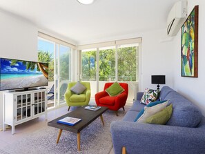 A living room with a television, two armchairs, a sofa, a coffee table, and a large window with a view of greenery. - Portofino 4 Hastings Street 59 (Noosa Heads)