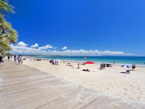 Una playa con un muelle de madera, gente caminando y sombrillas de playa.