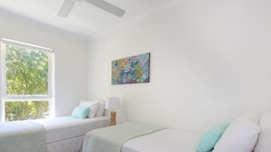 A room with two beds, a ceiling fan, a window with a view of trees, and a colorful abstract painting on the wall.