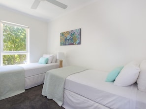 Una habitación con dos camas, un ventilador de techo, un ventanal con vistas a árboles y un cuadro abstracto de colores en la pared.