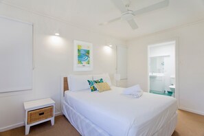 Habitación de hotel con cama, mesita de noche, cuadro enmarco, ventilador de techo y baño visible a través de una puerta abierta.