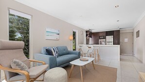 A modern living room with a sofa, armchair, and ottoman, a dining area with a table and chairs, and a kitchen with appliances.