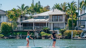 Two people paddleboarding on a calm body of water with a residential building in the background.