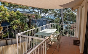 A balcony with a table and chairs, surrounded by a white railing, overlooking a residential area with trees and houses.