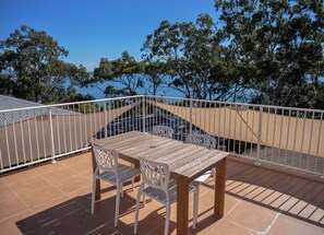 A terrace with a table and chairs, a white railing, and a view of trees and a body of water.