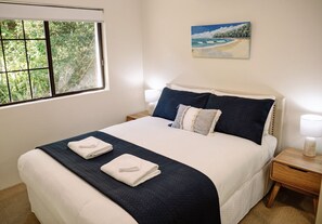 Un lit bien fait, recouvert d’une couvre-lit bleu foncé, avec deux oreillers et deux serviettes pliées. Une fenêtre offre une vue sur la verdure, et un tableau représentant une scène de plage est accroché au mur.