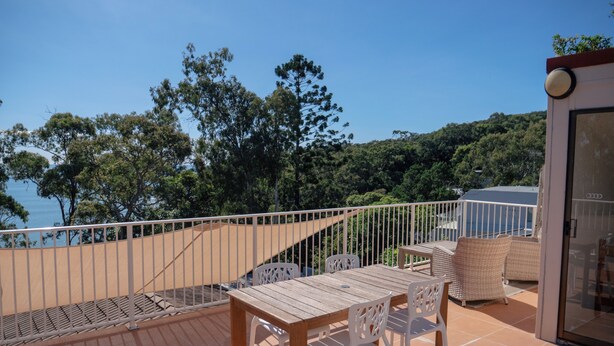 A terrace with a wooden table and chairs, a view of the sea, and a glass door leading to an interior space. - Costa Del Sol 3 Park Road 22 (Noosa Heads)