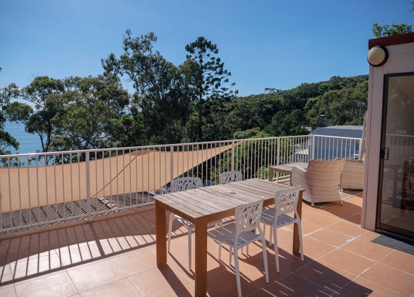 A terrace with a wooden table and chairs, a view of the sea, and a glass door leading to an interior space.