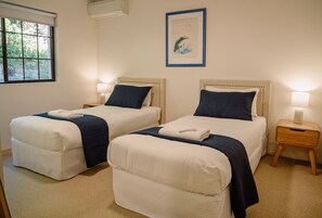 A hotel room with two beds, a window, an air conditioning unit, a framed picture on the wall, and a bedside table with a lamp.
