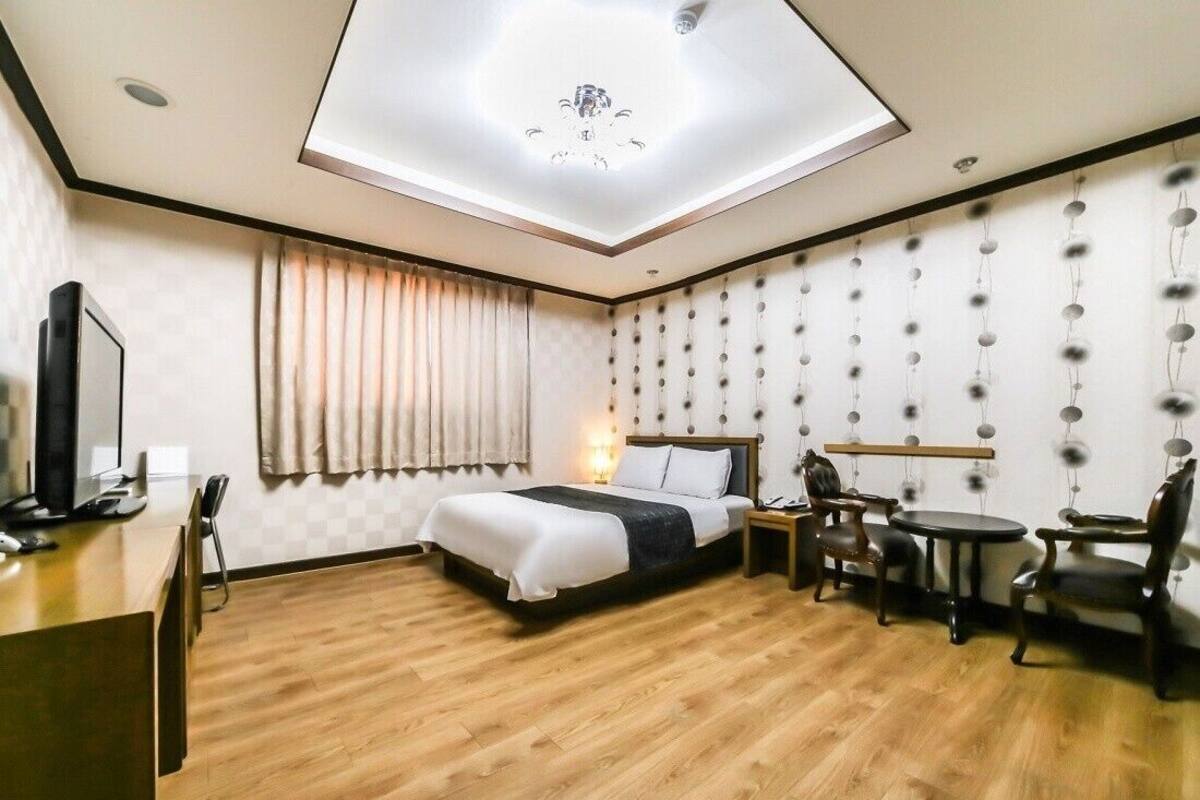 A hotel room with a bed, a desk, a chair, a small table, a TV, and a ceiling light.
