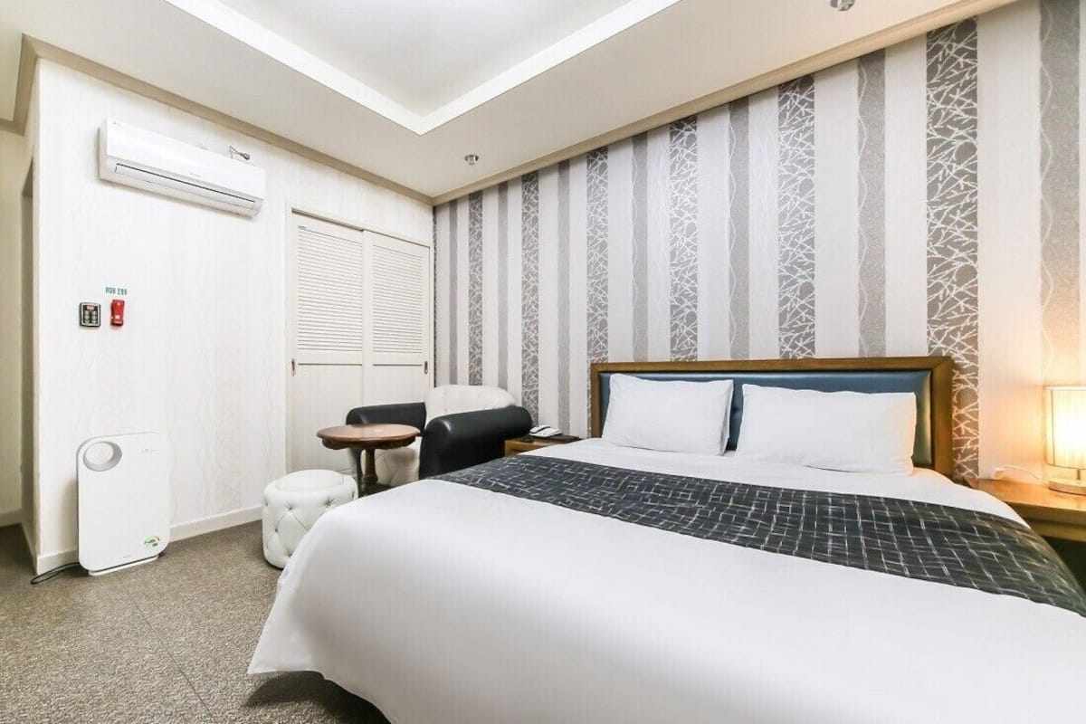 A hotel room with a large bed, a small table, a chair, an air conditioning unit, and a window with blinds.