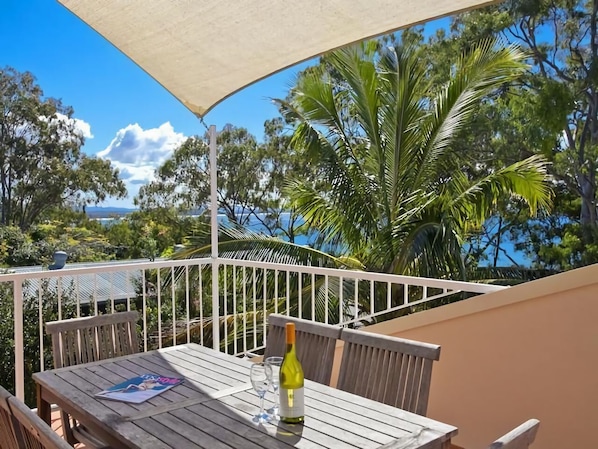 A wooden table with a bottle of wine and a glass, a white umbrella, and a white railing on a balcony with a view of trees and the sea.