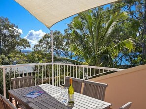 Une table en bois avec une bouteille de vin et un verre, un parasol blanc et une balustrade blanche sur un balcon d’où l’on peut admirer les arbres et la mer.
