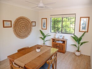 Un comedor con mesa de madera, sillas, un tapiz tejido y plantas en macetas.
