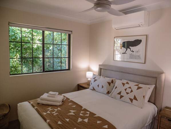 Habitación de hotel con cama, ventana con vistas a zonas verdes, ventilador de techo y un cuadro en la pared.