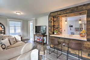 Un salon moderne avec un canapé, un téléviseur et un coin cuisine doté d’un mur en briques et de tabourets de bar.