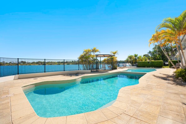 A swimming pool with a curved design, surrounded by a tiled deck and a metal fence.