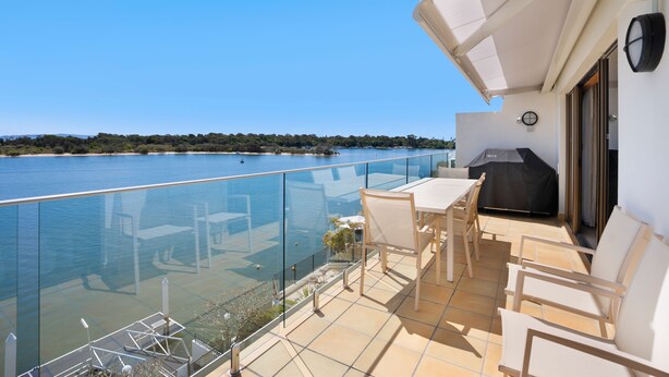 A balcony with a glass barrier overlooking a body of water, a table and chairs, and a grill area.