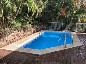 A rectangular swimming pool with a stone wall and metal railing, surrounded by a wooden deck and tropical vegetation.