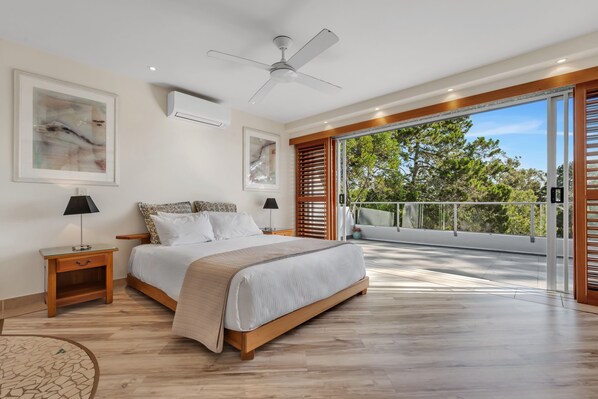 A bedroom with a large bed, a ceiling fan, and a view of trees through a sliding glass door.