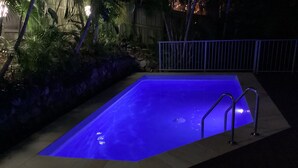 Une piscine rectangulaire dont l’intérieur est illuminé de lumière bleue, entourée d’un mur de pierre et d’une clôture en métal.