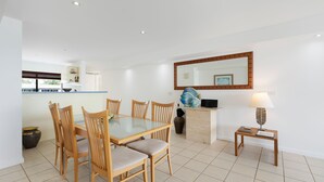 A dining area with a glass-top table and wooden chairs, a kitchen area with a counter and cabinets, and a small side table with a lamp. - Aqua Linea 3 Noosa Parade 82 (Noosa Heads)