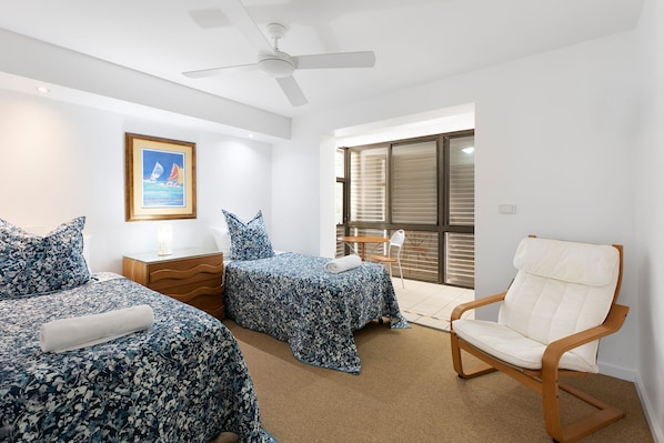 A hotel room with two beds, a wooden chair, a ceiling fan, a framed picture, and a window with blinds.