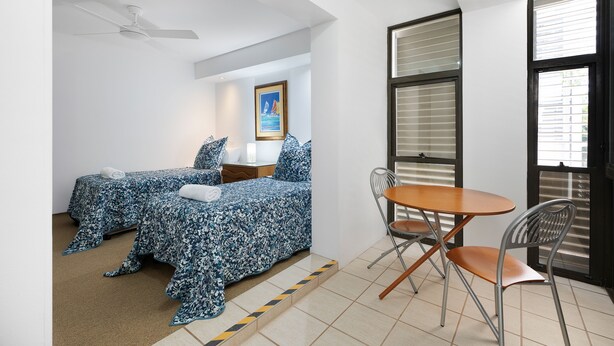 A bedroom with two beds, a ceiling fan, a window with blinds, and a small table with chairs. - Aqua Linea 3 Noosa Parade 82 (Noosa Heads)