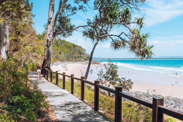 Una passeggiata costiera con ringhiera in legno che conduce a una spiaggia con acque cristalline e un paesaggio collinare sullo sfondo.