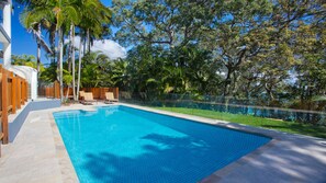 A swimming pool with a tiled deck, surrounded by palm trees and a wooden fence.