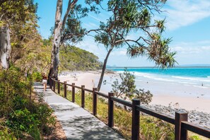 Una passeggiata costiera con ringhiera in legno, che conduce a una spiaggia con acque blu cristalline e un paesaggio collinare sullo sfondo.