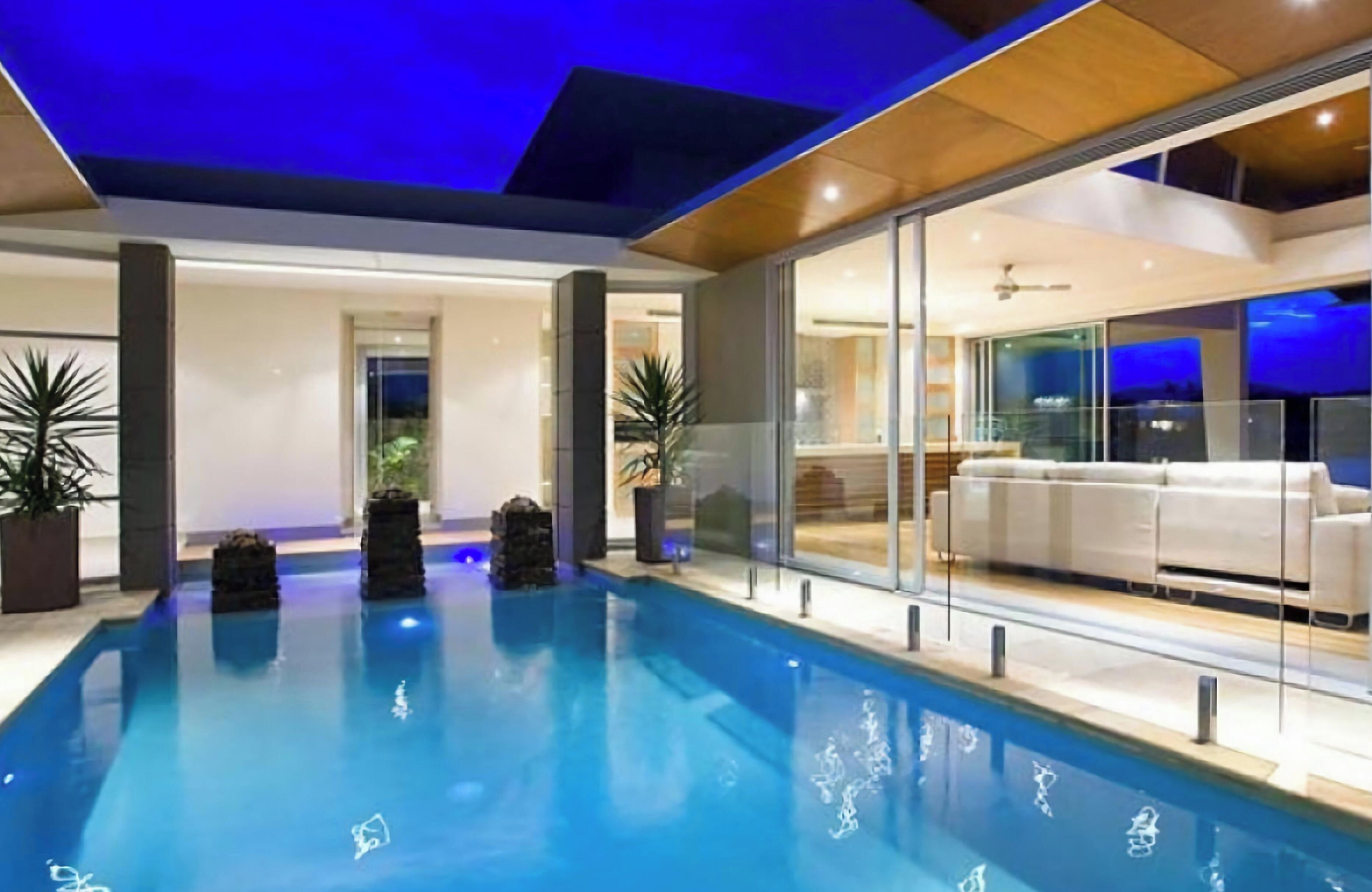 A modern indoor swimming pool with clear glass walls, a white sofa, and potted plants.