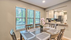 Un coin repas avec une table dressée pour quatre personnes, une orchidée en pot et une vue sur la cuisine à travers une fenêtre munie de stores.