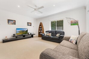 A modern living room with a flat-screen TV, a black leather sofa, a wooden bookshelf, and a ceiling fan. - Treasure Cove 6 (Noosaville)