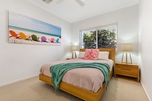 A bedroom with a bed, a nightstand, a window with blinds, a wall-mounted painting of beach chairs, and a ceiling fan. - Treasure Cove 6 (Noosaville)