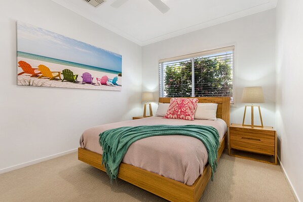 Une chambre à coucher comprenant un lit, une table de chevet, une fenêtre avec des stores, un tableau représentant des chaises de plage accroché au mur, et un ventilateur de plafond.