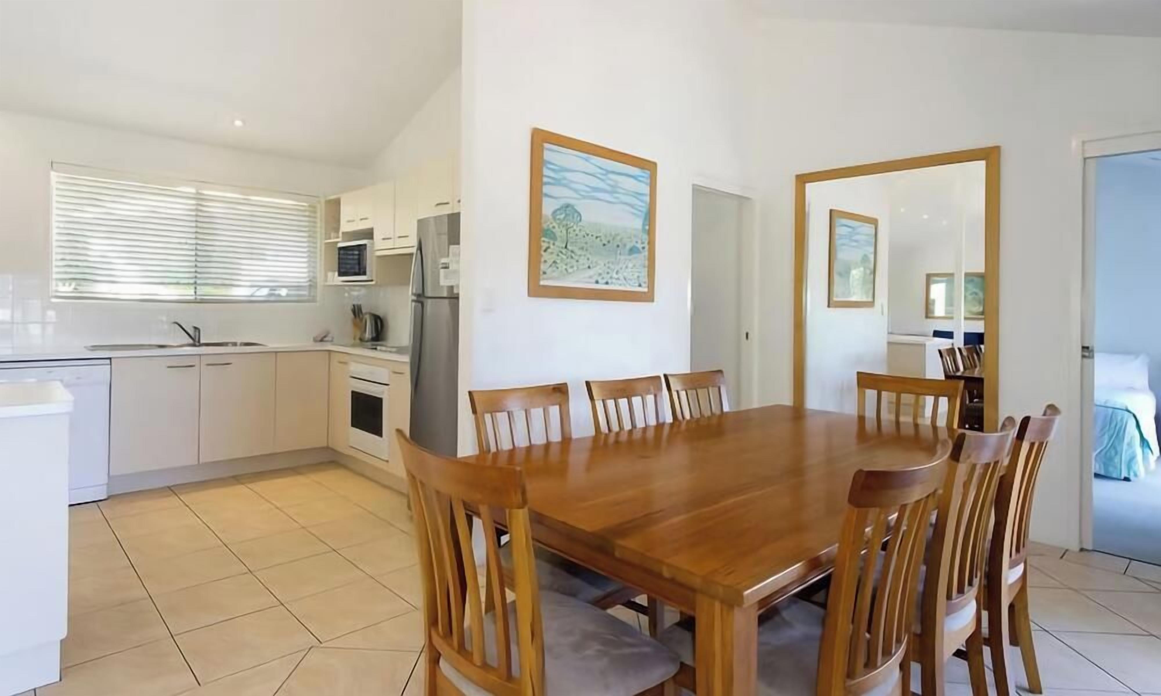 A modern kitchen with white cabinets, a wooden dining table, and chairs. There is a window with blinds, a refrigerator, and a microwave.