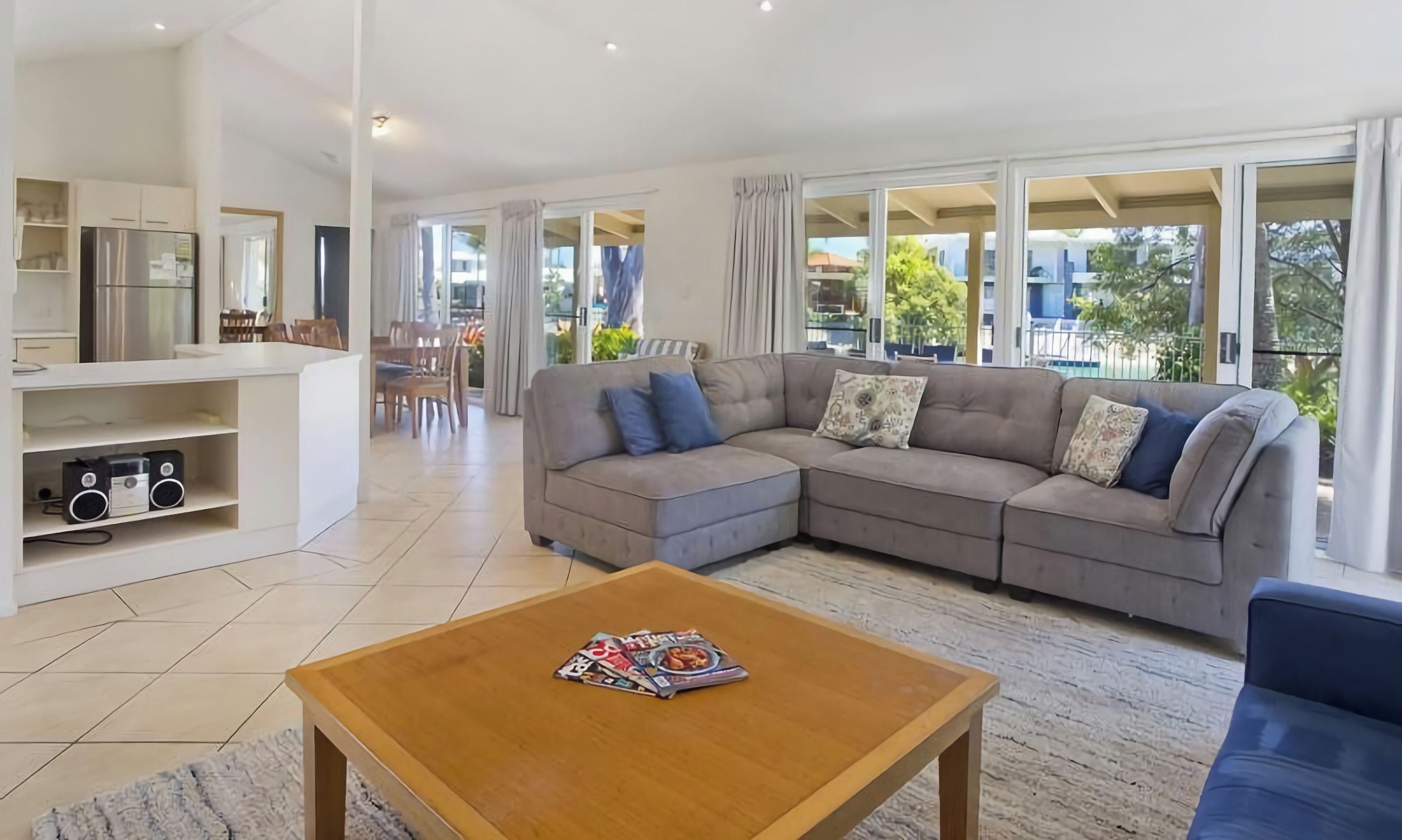 A modern living room with a sectional sofa, a coffee table with magazines, a kitchen area with a refrigerator, and large windows offering a view of the outdoors.