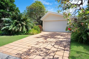 Une maison avec une allée dallée menant à un garage, entourée d’une végétation luxuriante et de palmiers.