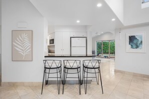 A modern kitchen with a central island, three bar stools, and artwork on the walls. - Witta Circle 24 (Noosa Heads)