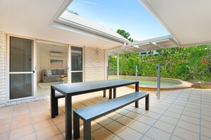 Une terrasse avec une table et un banc, une porte-fenêtre coulissante donnant sur un espace de vie et un espace extérieur couvert agrémenté de verdure.