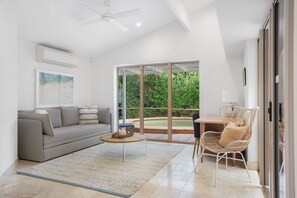 Un salon moderne avec un canapé, une table basse et un fauteuil en osier, de grandes portes-fenêtres coulissantes donnant sur un jardin.