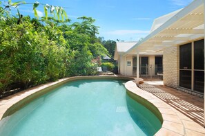 Une piscine dont le bord est revêtu de carrelage, entourée d’une végétation luxuriante et dotée d’un patio couvert.