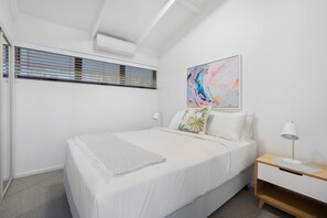 A modern bedroom with a bed, a nightstand, a lamp, a painting, and a window with blinds. - Witta Circle 24 (Noosa Heads)