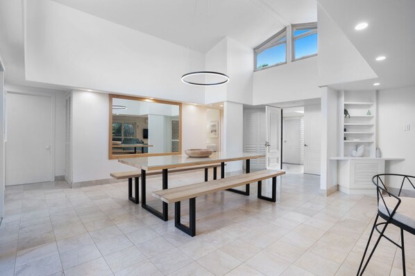 Un interior espacioso y moderno con techo alto, un espejo grande, una zona de comedor con bancos y vistas al exterior a través de una ventana.