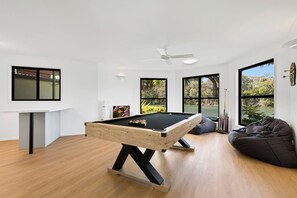 Una mesa de billar moderna en interior con estructura de madera, ventilador de techo y vistas a un lago a través de amplios ventanales.