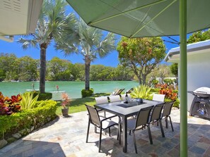 Un patio exterior con mesa y sillas, una parrilla y vista a un lago con palmeras.