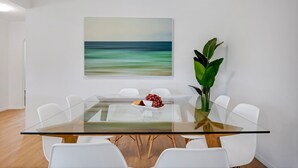 A modern dining room with a glass table, white chairs, a painting of a beach, and a large green plant. - Wyuna Drive 53 (Noosaville)